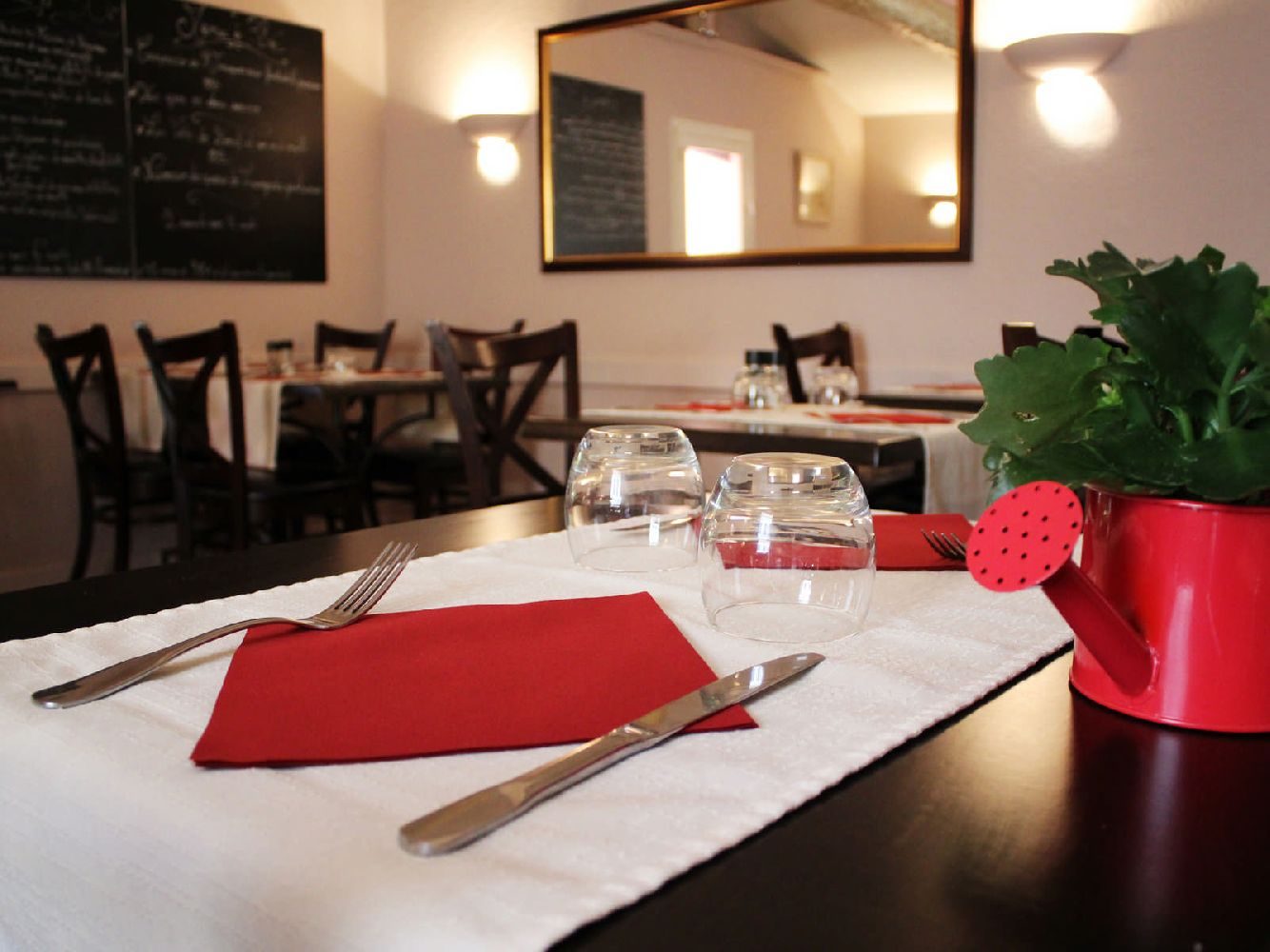 Une table dressée : chemin de table blanc, serviette rouge, verres et petit arrosoir rouge garni de verdure