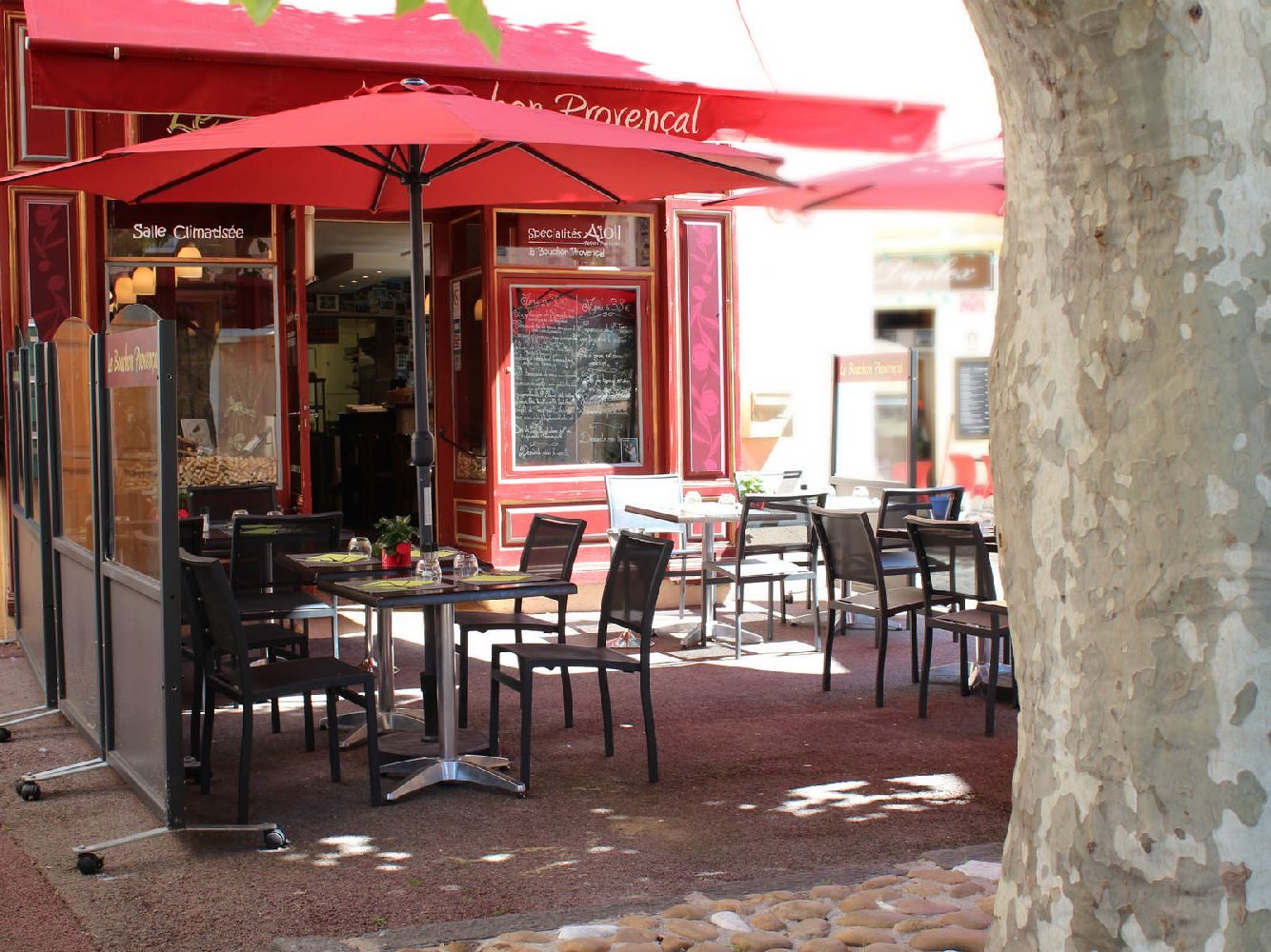 La terrasse sous un grand platane et des parasols rouges, tables dressées devant la façade rouge